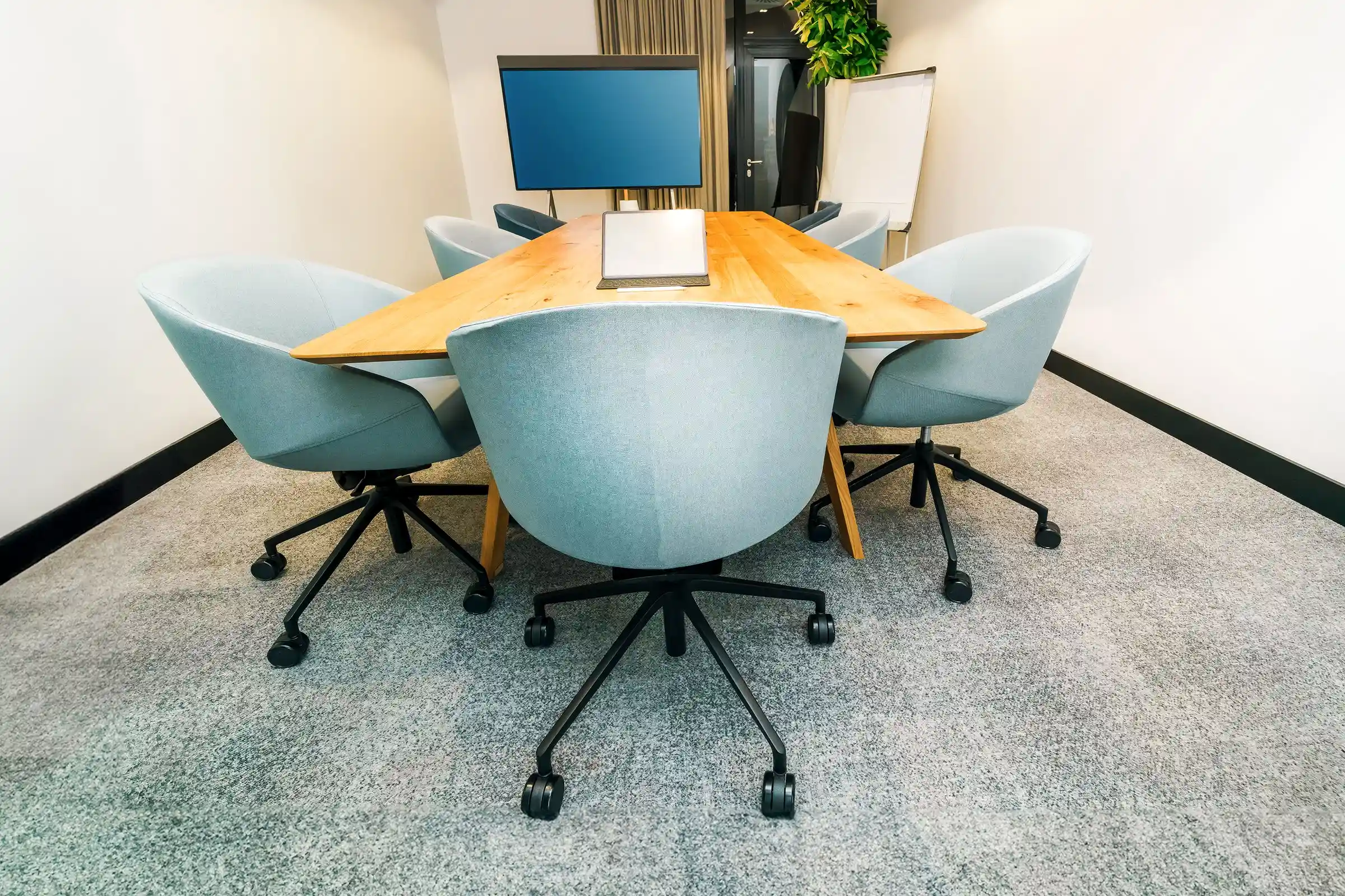 An empty meeting room with pale blue chairs around a wooden table, a monitor behind.