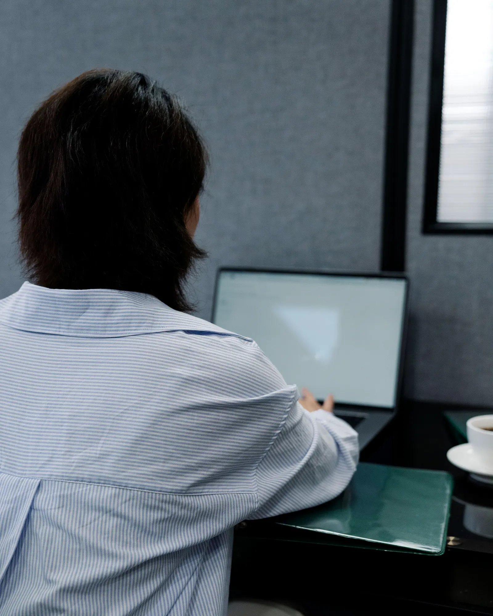 A person seen from behind at a desk, working at a laptop, a coffee cup beside it.