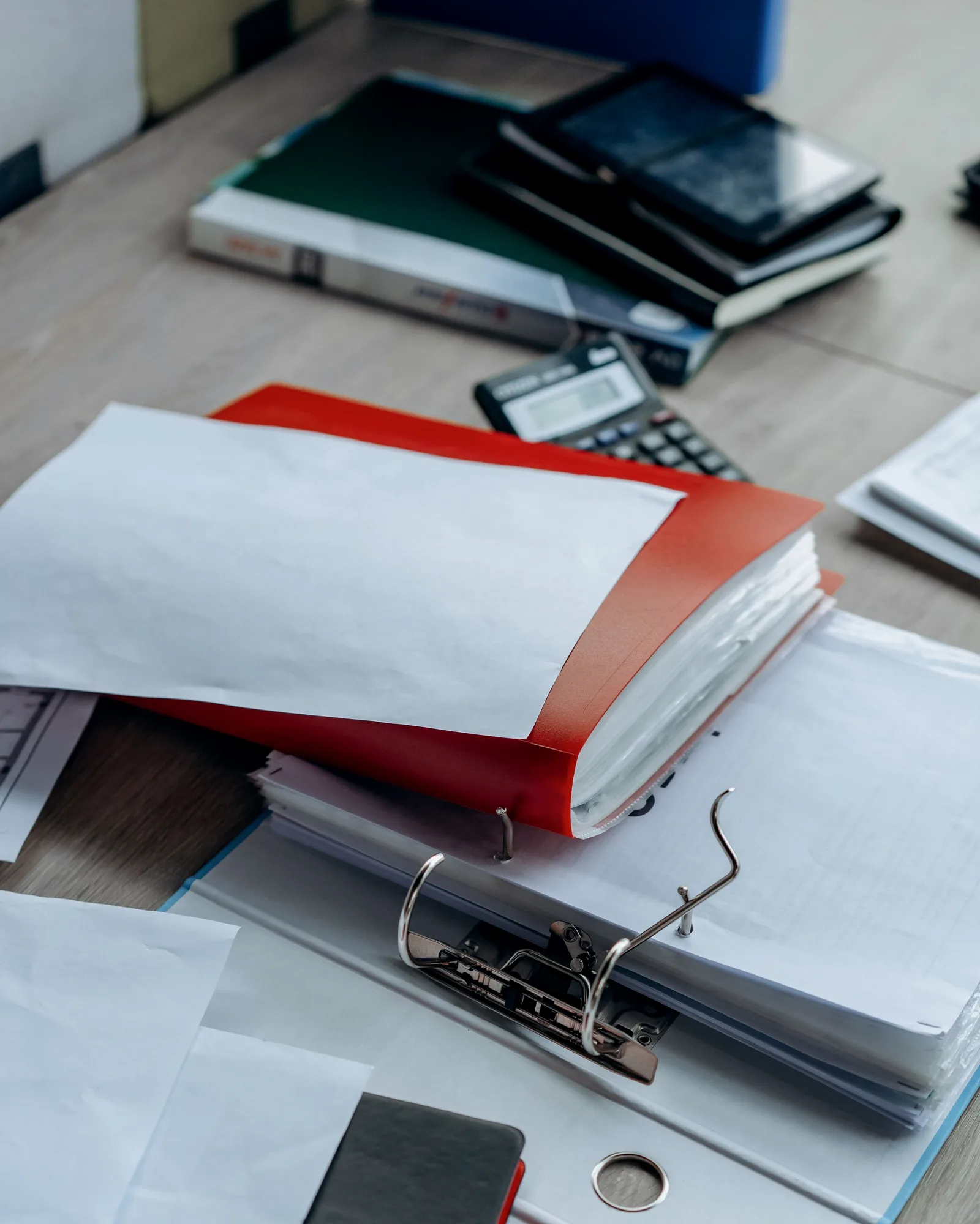 A red ring binder and papers on a desk, a stack of binders and a calculator behind.