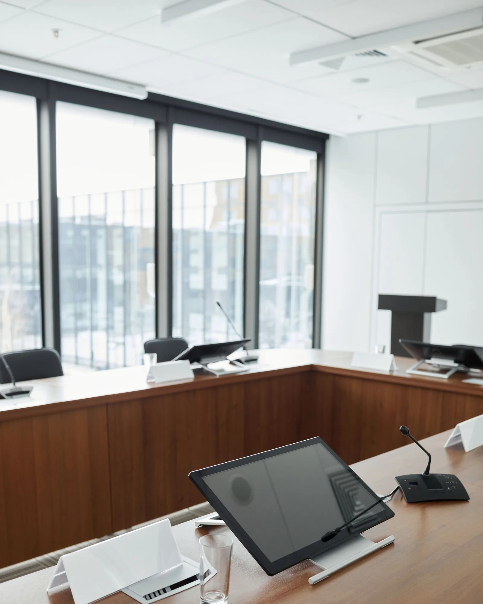 An empty boardroom with a wood-veneer table, a microphone and a tablet, windows behind.