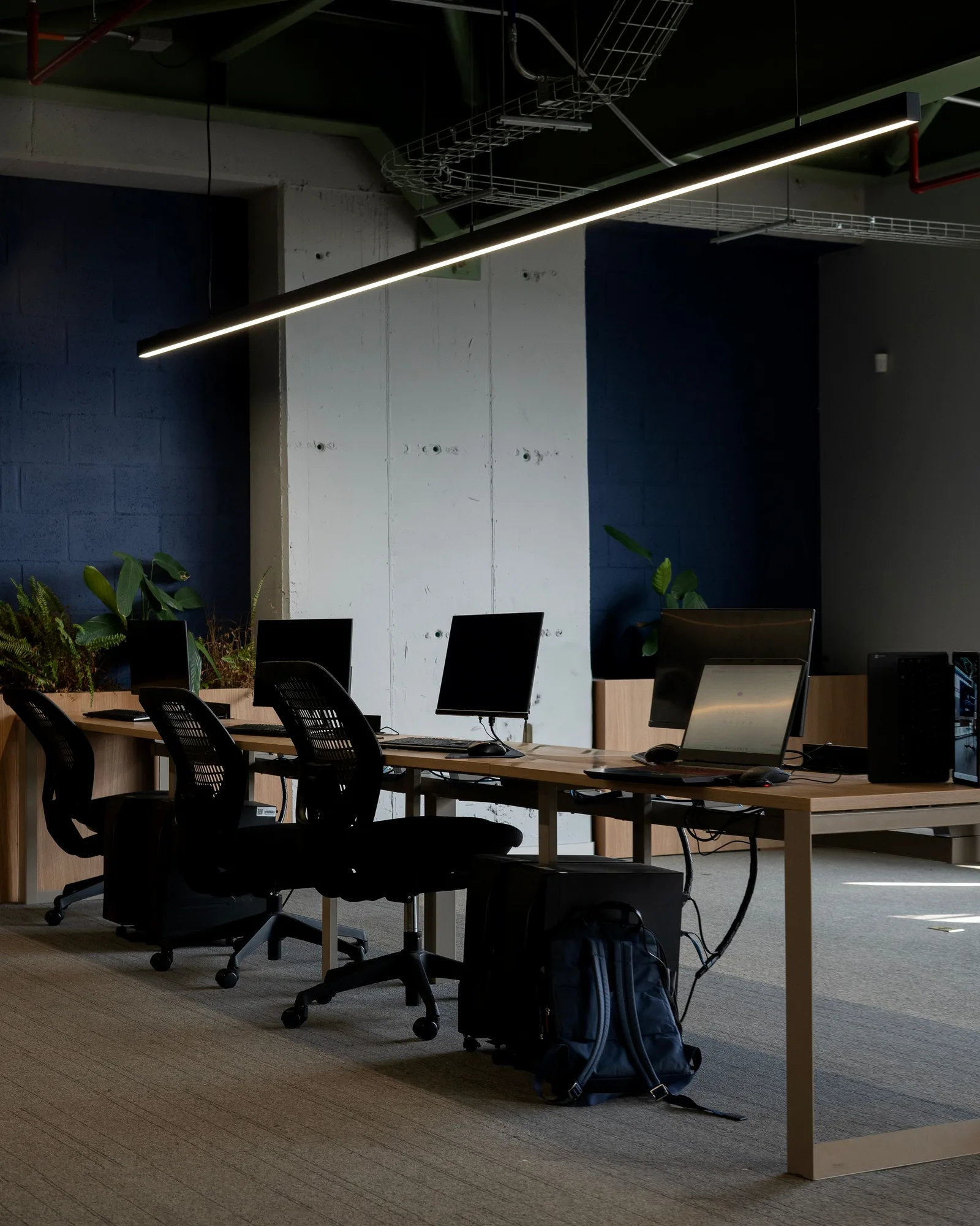 An empty office workstation row with monitors and a laptop, under a long pendant light.